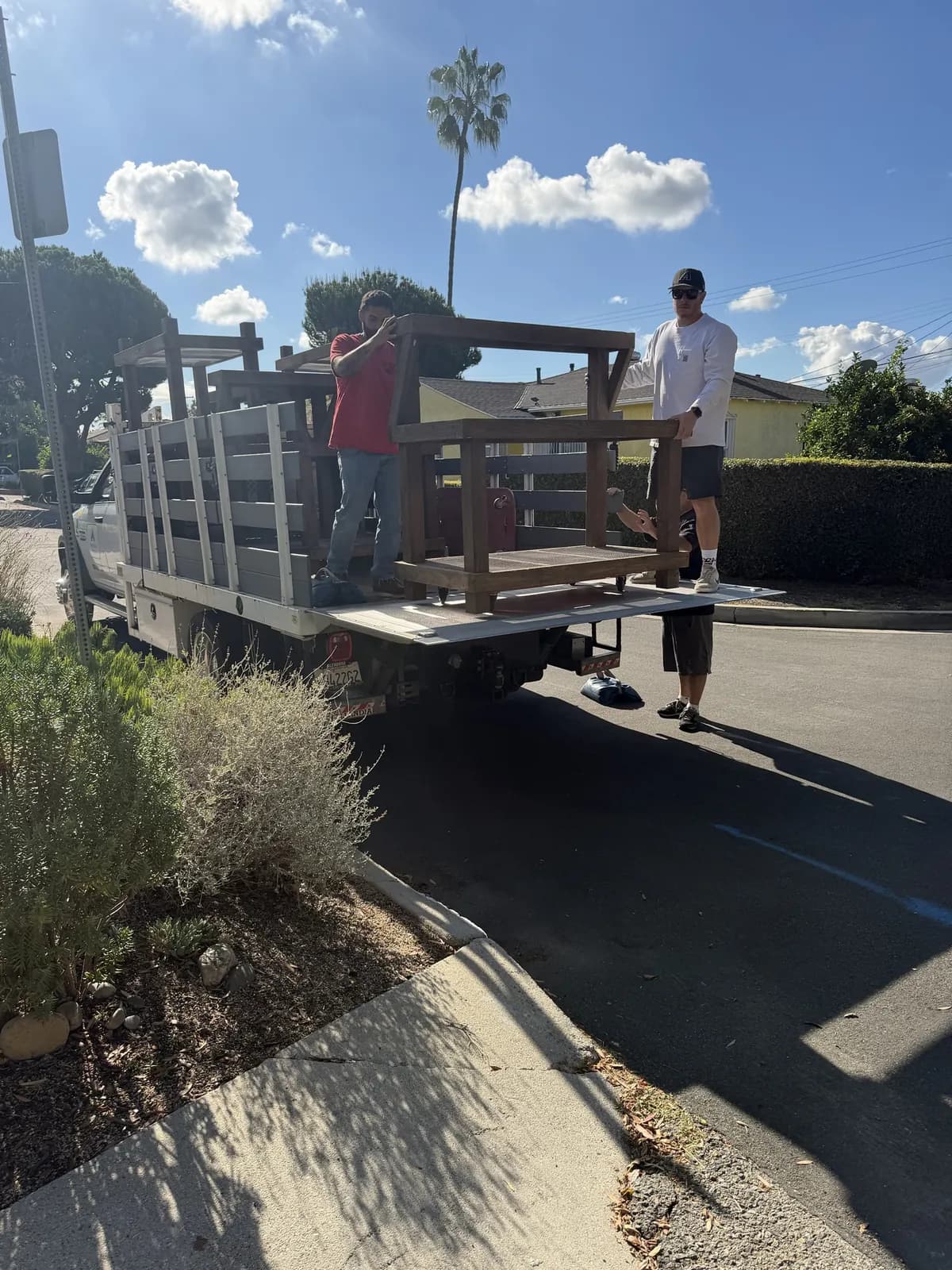 Two people easing a bench module off a stake-bed truck's liftgate at the curb
