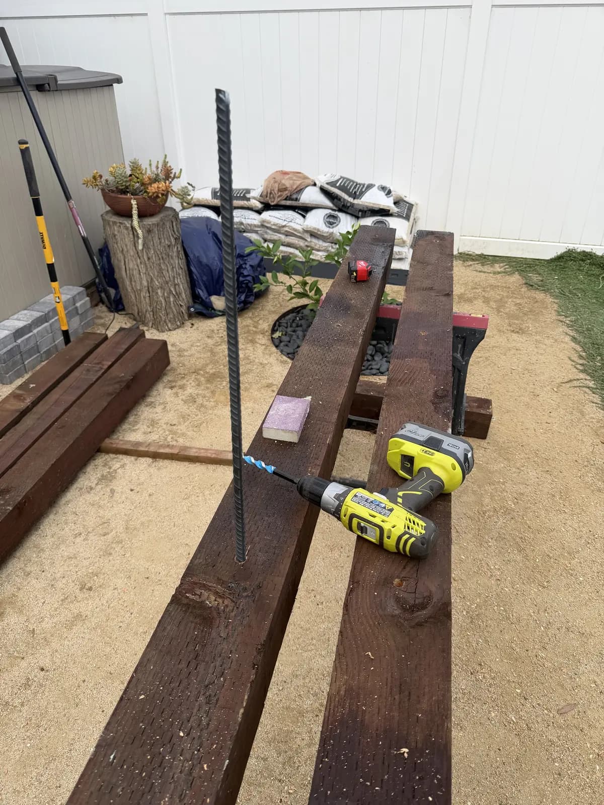 A length of rebar test-fit upright in a hole drilled through a 6×6, the cordless drill and auger bit beside it
