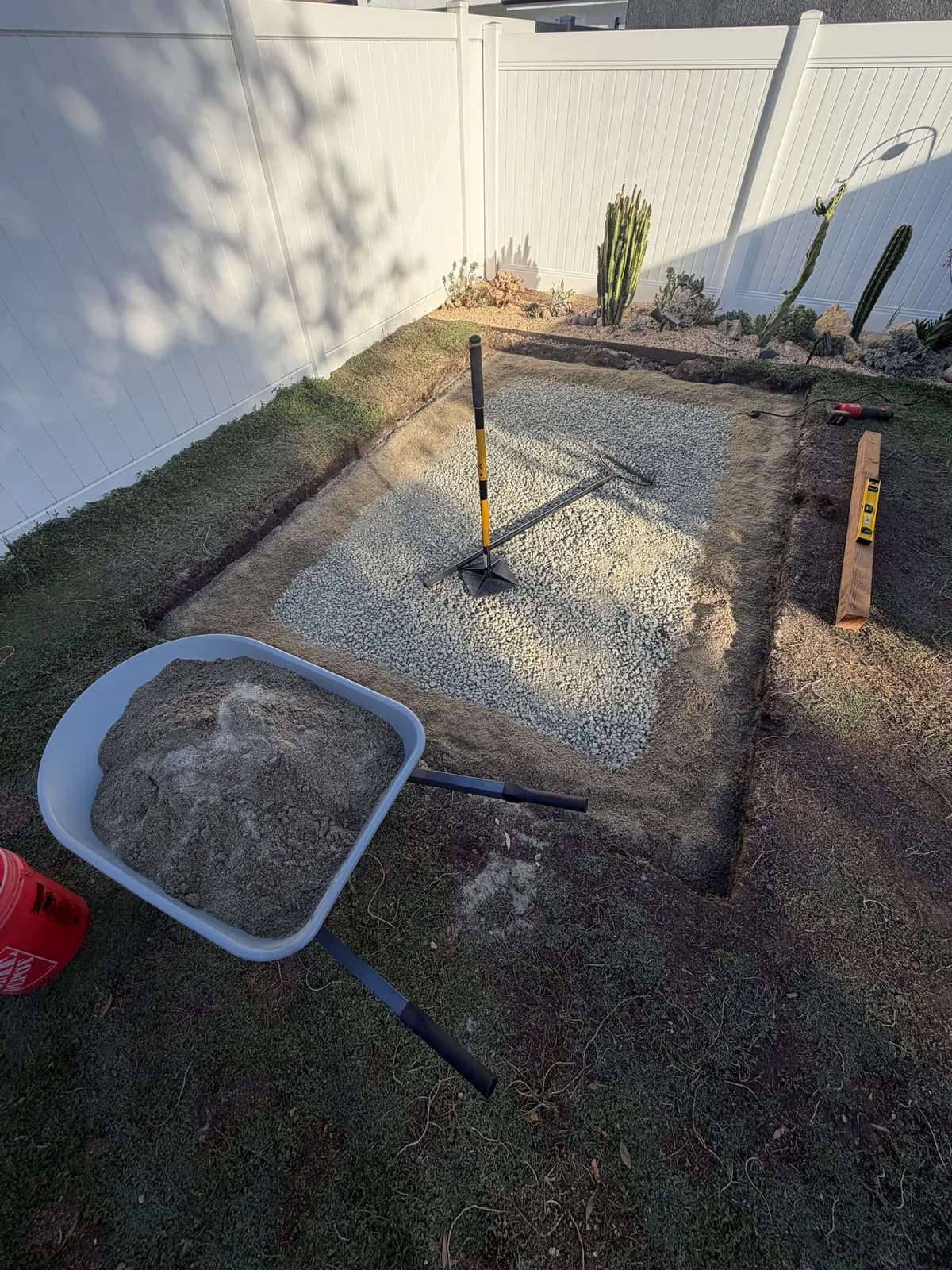 A wheelbarrow of sand beside the pad, sand spread around the edge of the gravel and a tamper in the middle