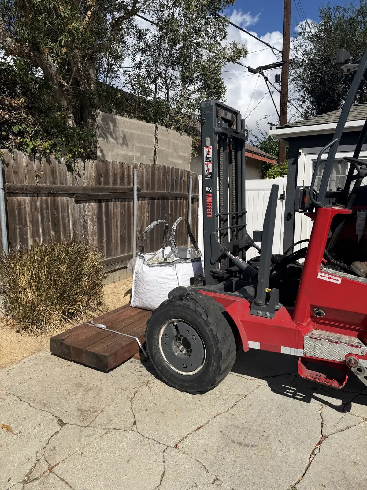 A strapped bundle of 6×6 timbers dropped by a forklift at the curb, next to a bulk bag of gravel