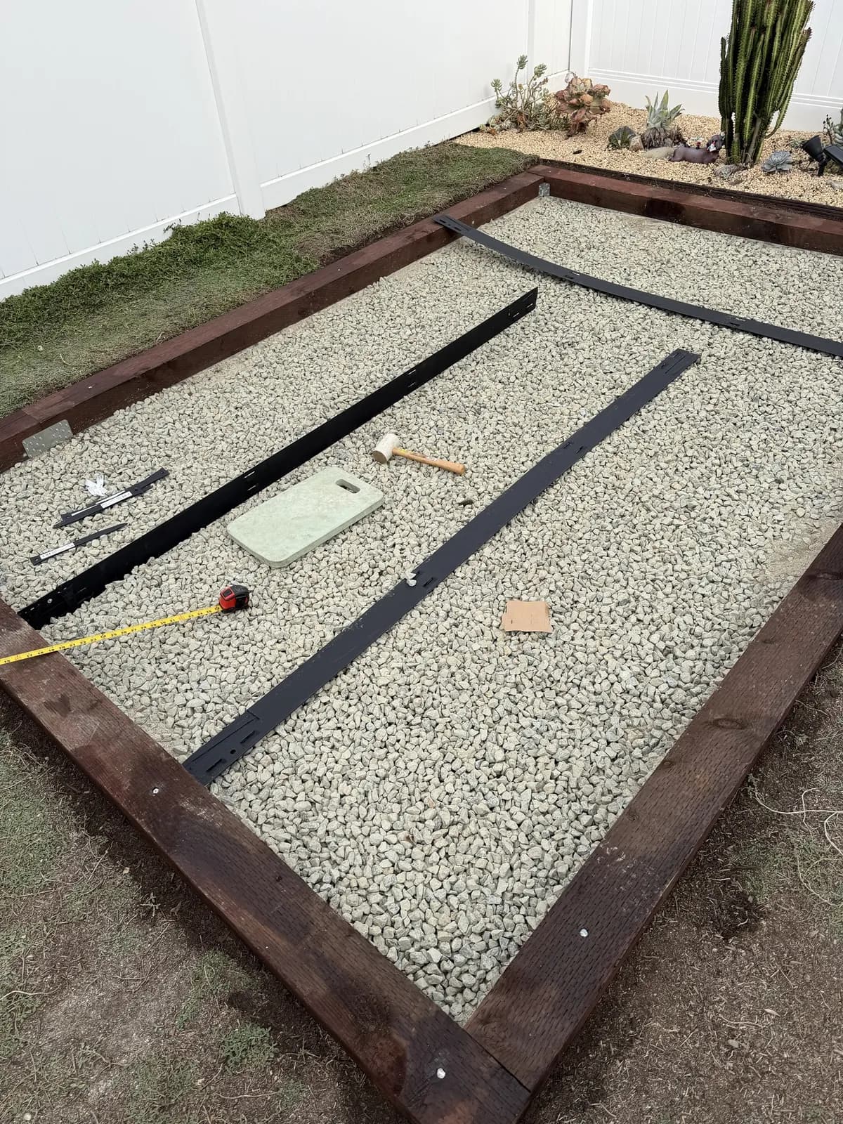 Black steel edging strips laid out on the gravel with a tape measure, rubber mallet, stakes, and a kneeling pad